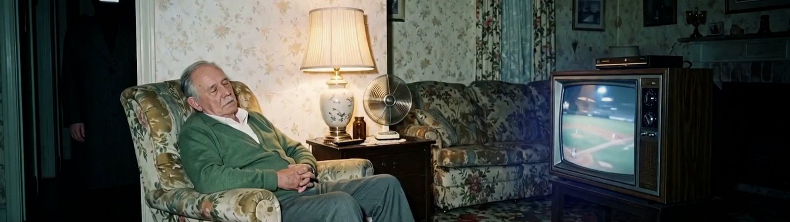 A grandfather asleep in a floral armchair beside a lamp and a fan, a ball game playing on an old wooden television