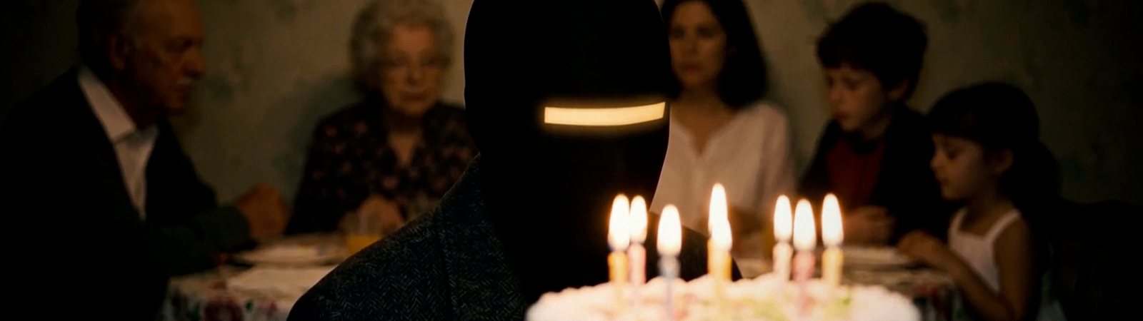 Close on the faceless figure at the birthday table, its bar of light glowing warm amber over lit candles